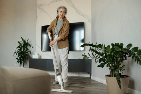 Elderly woman vacuuming living room with modern decor, standing between two potted plants and TV on wall. She appears focused while performing household choreの写真素材