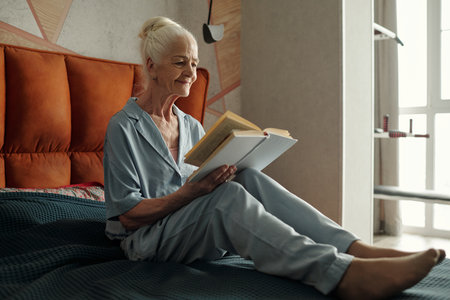 Elderly woman dressed in pajamas sitting comfortably on bed reading a book in cozy bedroom. Natural light coming through window illuminates scene, creating calm atmosphereの写真素材