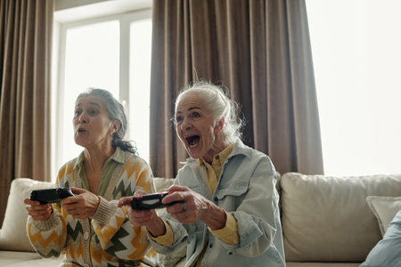 Elderly women enjoying gaming on sofa, holding controllers and expressing excitement and joy,...