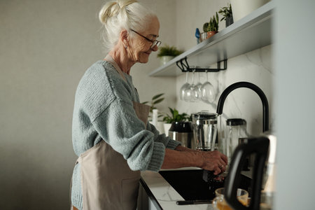 Senior woman washing dishes in modern kitchen with various kitchen gadgets on counter. Light-filled...