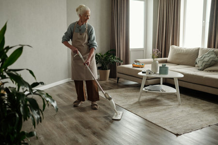 Senior woman vacuuming living room floor with modern decor, including sofa, coffee table, and curtains. Light entering through big windows illuminating the spaceの写真素材