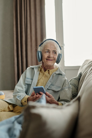 Elderly woman enjoying music with headphones while holding smartphone and sitting on couch, looking away from camera, in cozy, well-lit living room, looking content and relaxedの写真素材