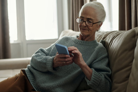 Elderly woman with glasses enjoying free time on comfortable couch while engaging with smartphone....