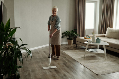 Elderly woman vacuuming wooden floor in living room with modern furniture and large windows bringing natural light. Home neatly arranged with sofa, coffee table, and potted plantsの写真素材