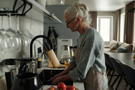Elderly woman standing at kitchen sink, washing vegetables with focus and determination. Modern kitchen setting with various kitchen utensils and appliances visibleの写真素材