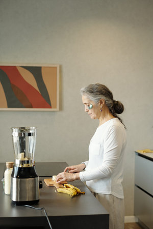 Elderly woman standing in modern kitchen, blending ingredients with a blender. Fresh bananas and...