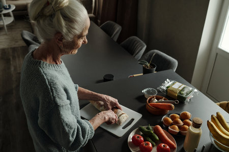 Elderly woman slicing bread on a cutting board with fresh vegetables around her, engaging in cooking activity in a serene kitchen environmentの写真素材
