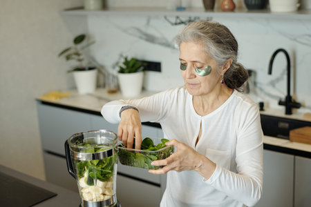 Elderly woman blending fresh spinach and bananas in modern kitchen for smoothie preparation,...