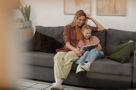 Candid portrait of smiling mother and daughter watching videos online together and looking at tablet screen while sitting on soft comfortable couch copy spaceの写真素材