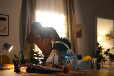 African American man leaning over table, helping a child focused on homework, illuminated by indoor lighting with a cozy living room in backgroundの写真素材