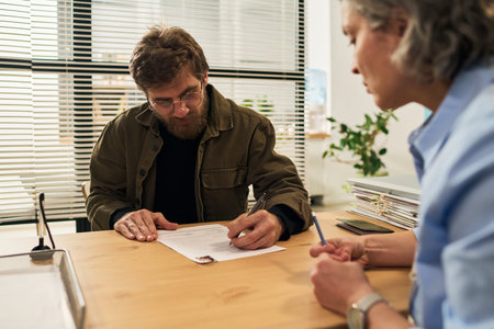 Middle aged Caucasian man sitting at desk signing paperwork with middle aged Caucasian female social...
