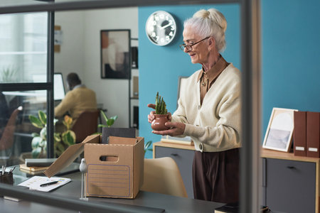 Senior Caucasian woman standing at desk holding small potted plant, packing personal belongings into cardboard box, preparing for retirement in modern office settingの写真素材