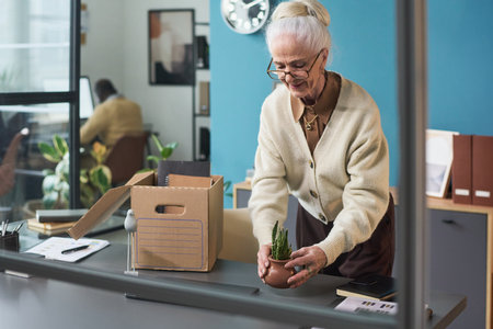 Senior Caucasian woman unpacking cardboard box and arranging small potted plant on desk in modern office studio, wearing glasses and smiling while standing near windowの写真素材