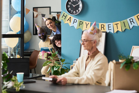 Senior woman sitting at desk while three colleagues wearing party hats smiling and peeking from behind wall during retirement celebrationの写真素材