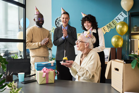 Senior Caucasian woman sitting at desk holding cupcake with candle while young adults standing behind her clapping and smiling during retirement celebration in studioの写真素材