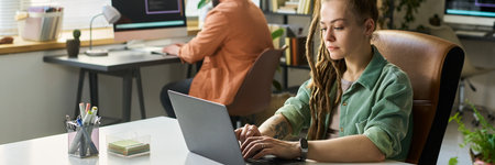 Young adult Caucasian woman developing neural network on laptop, sitting at desk in modern office, multiethnic colleague working at computer in background, technology workspaceの写真素材
