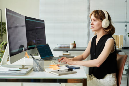 Caucasian young adult woman wearing headphones working on neural network development at desk using dual monitors coding and analyzing data in modern office settingの写真素材