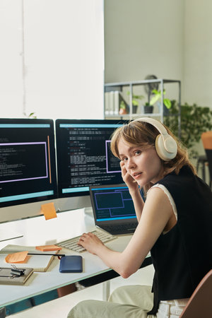 Portrait of young adult Caucasian woman wearing headphones sitting at desk coding neural network on computer monitors, looking at camera, using laptop and taking notesの写真素材