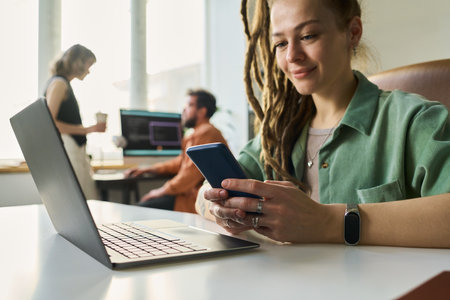 Caucasian young adult woman sitting at desk using smartphone and laptop, smiling while working on neural network development, two colleagues collaborating in background with computer screensの写真素材