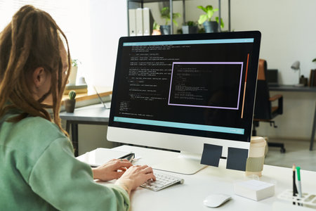 Young adult Caucasian woman sitting at desk working on neural network development coding on large desktop computer screen in modern officeの写真素材