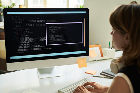 Caucasian young adult woman developing neural network code on desktop computer, sitting at desk, focusing on programming interface with lines of code visible on large monitorの写真素材