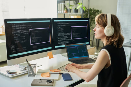 Caucasian young adult woman wearing headphones working on neural network development using dual monitors displaying code in modern office setting, typing on keyboard, focusing on screenの写真素材