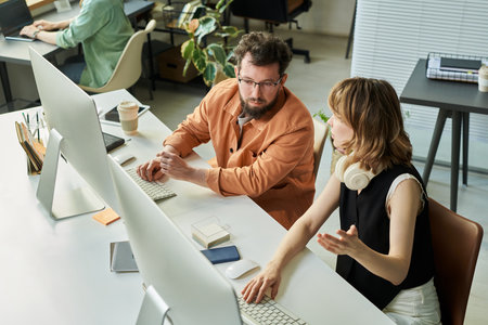 Caucasian middle aged man and Caucasian young adult woman collaborating at workstation, discussing neural network development while using desktop computers in modern office settingの写真素材