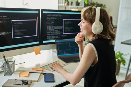 Caucasian young adult woman wearing headphones working on neural network development at computer workstation, smiling while coding on multiple monitors with programming interface visibleの写真素材