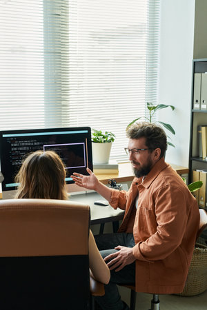 Caucasian middle aged man explaining neural network code to Caucasian young adult woman sitting at desk using large computer monitor in modern office settingの写真素材