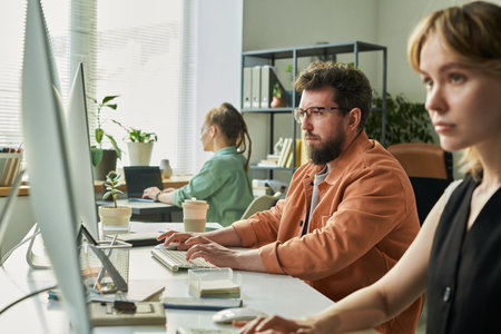 Caucasian young adult man and Caucasian young adult woman working at computers in modern office, focused on neural network development, female colleague in background using laptopの写真素材