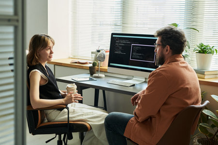 Woman holding coffee cup discussing neural network development with Caucasian middle aged man sitting at desk with computer monitor displaying code in modern officeの写真素材