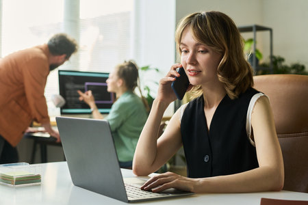 Caucasian young adult woman sitting at desk using laptop and talking on smartphone developing neural network project on computerの写真素材