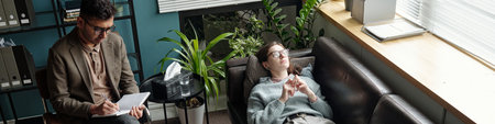Young man lying on sofa talking during therapy session with male therapist sitting nearby writing notes in notebook in modern office settingの写真素材