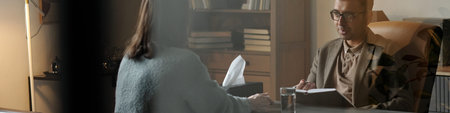 Man sitting at desk holding clipboard engaging in therapy session with woman sitting across table in...