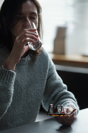 Woman sitting at table holding eyeglasses in one hand drinking water from glass with eyes closed suggesting therapy or self care routineの写真素材