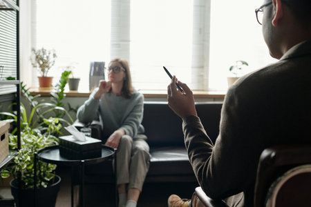 Woman sitting on sofa talking with man holding pen during therapy session in office setting with plants and window in backgroundの写真素材
