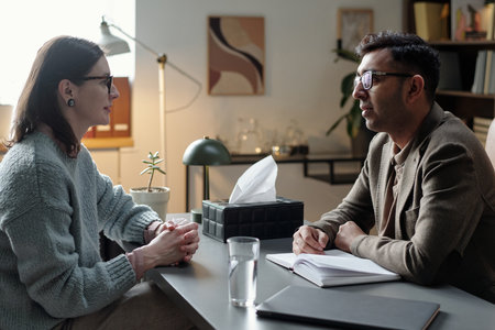 Woman sitting across from man taking notes during therapy session in office setting, both maintaining eye contact and engaged in conversationの写真素材