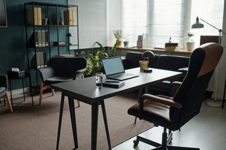 Empty modern therapy office showing desk with open laptop, notebook, glass of water and armchair, suggesting professional setting for counseling or psychological consultation sessionの写真素材