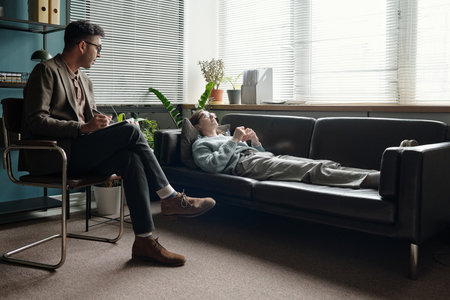 Woman lying on sofa talking to man therapist sitting with notebook listening attentively during therapy session in modern office with window blindsの写真素材
