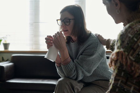 Woman sitting and holding tissue while crying, receiving comforting gesture from another woman during therapy session in office settingの写真素材