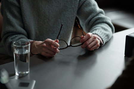 Woman holding eyeglasses with both hands sitting at table during therapy session, glass of water and...
