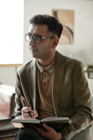 Man sitting with notebook and pen listening attentively during therapy session, wearing glasses and...