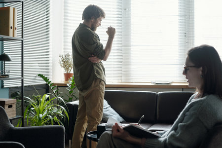 Man standing near window thinking during therapy session while woman therapist sitting on sofa writing notes in notebookの写真素材