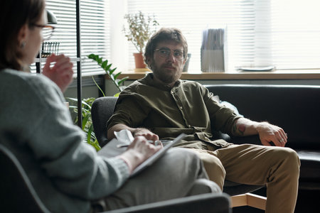 Man sitting on sofa engaging in therapy session with woman therapist holding clipboard, both facing each other in professional office settingの写真素材