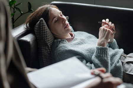 Woman lying on couch holding eyeglasses during therapy session, therapist with notepad listening attentively, mental health support conceptの写真素材