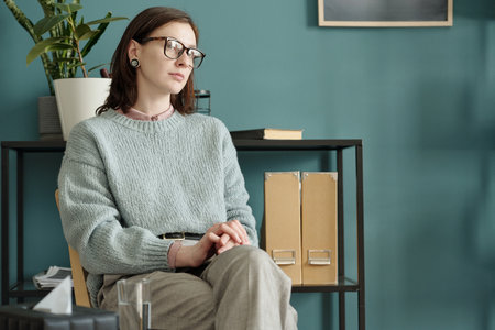 Woman sitting with hands clasped, wearing glasses, looking away thoughtfully during therapy session in modern office setting, plant and folders in backgroundの写真素材