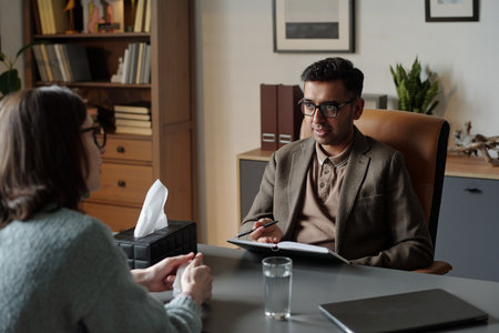 Man conducting therapy session with woman, therapist holding clipboard and pen, client sitting across desk, tissue box and glass of water visibleの写真素材