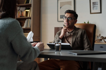 Man sitting at desk holding pen listening attentively to woman during therapy session in office...