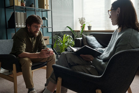 Man sitting with head down holding hands together while woman therapist listening and taking notes during therapy session in officeの写真素材