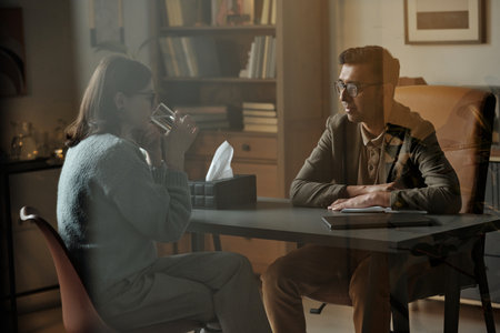 Woman sitting across from man wearing glasses, engaging in therapy session, man listening attentively with notebook on tableの写真素材